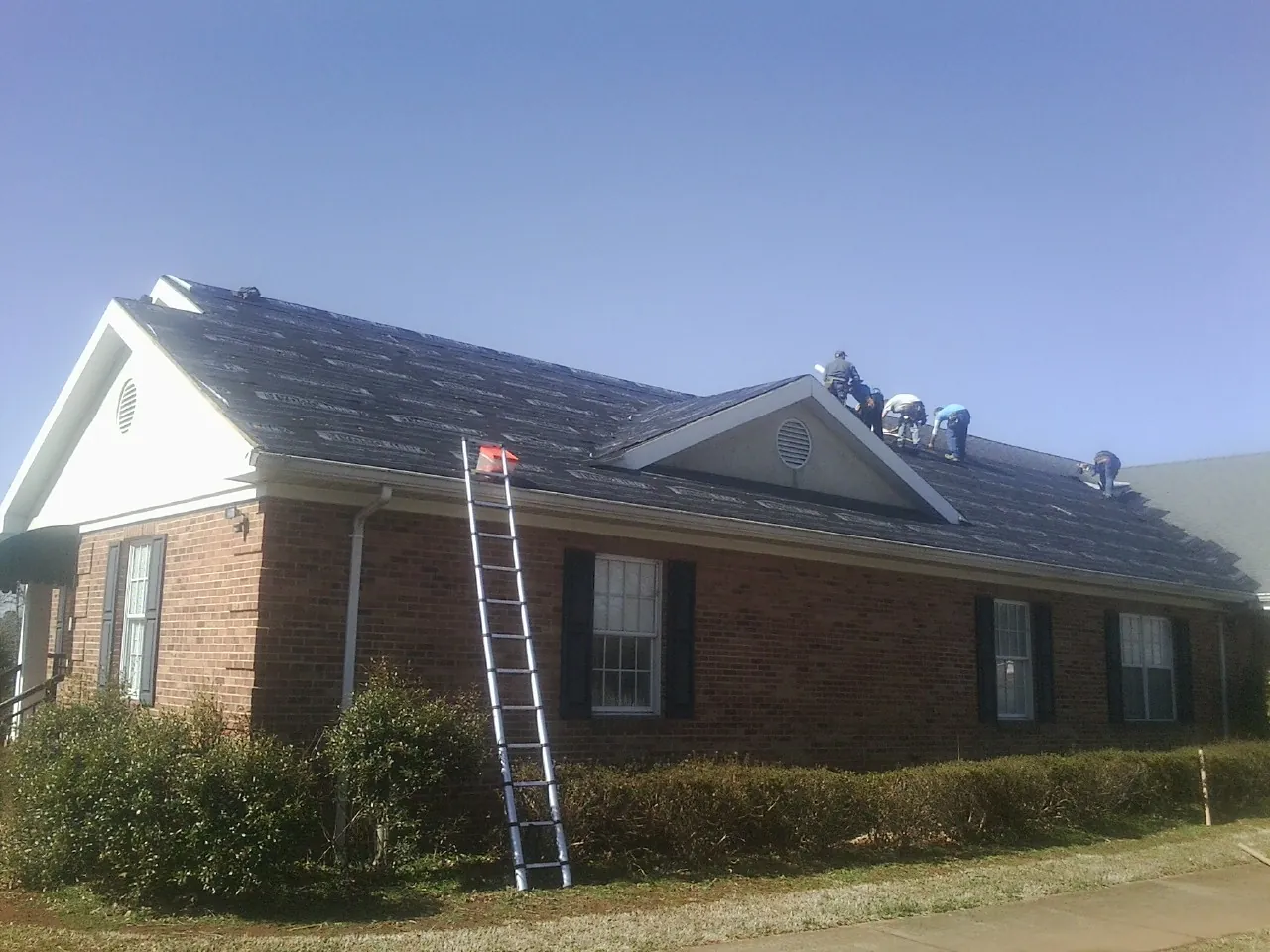 Crew tearing shingles off a brick ranch roof with a ladder set — Metal Roof Repair work by our crew in Buna