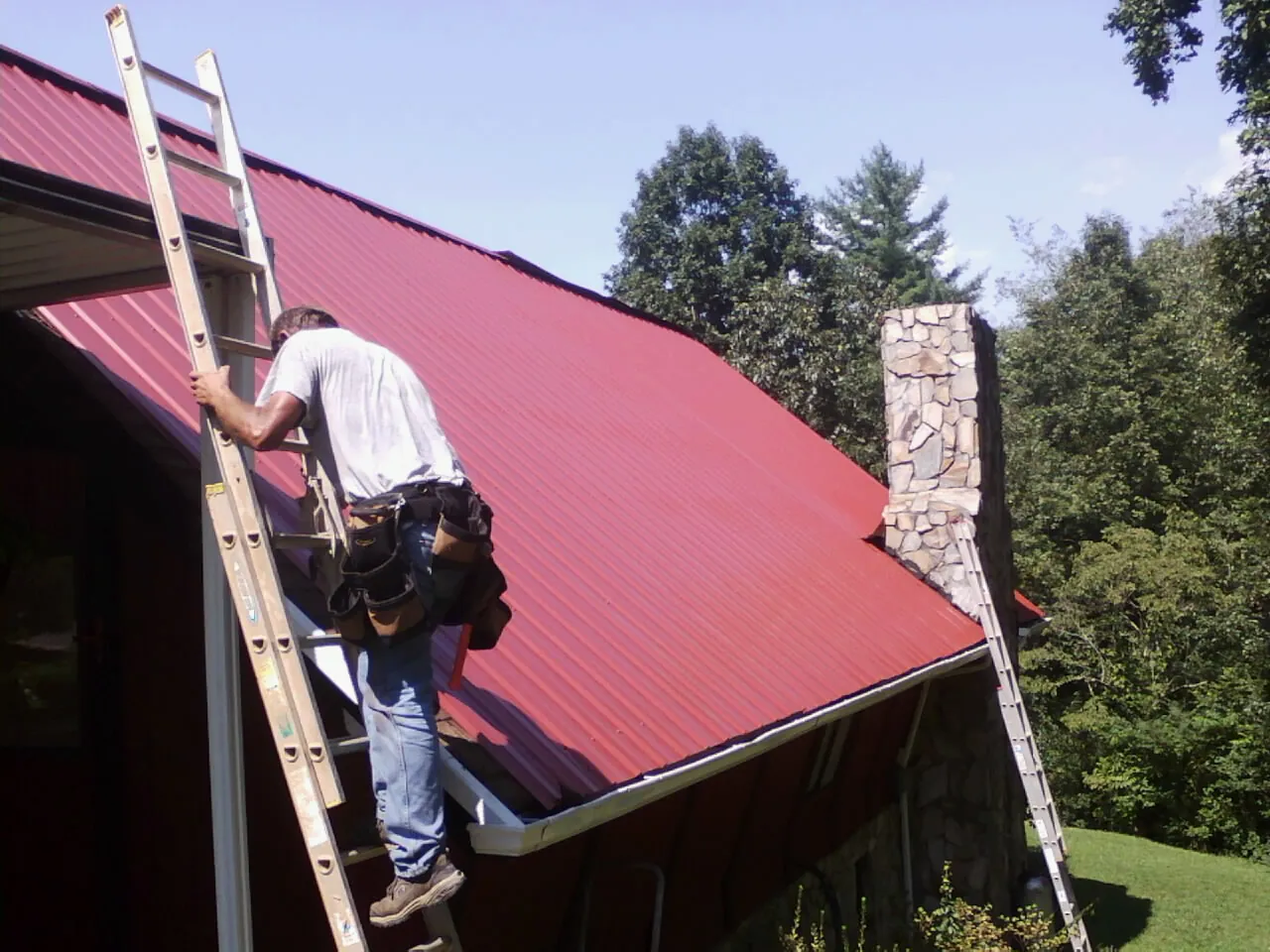 Roofer on a ladder beside a red metal roof and stone chimney — Soffit & Fascia Repair by Roofing Brothers in Buna